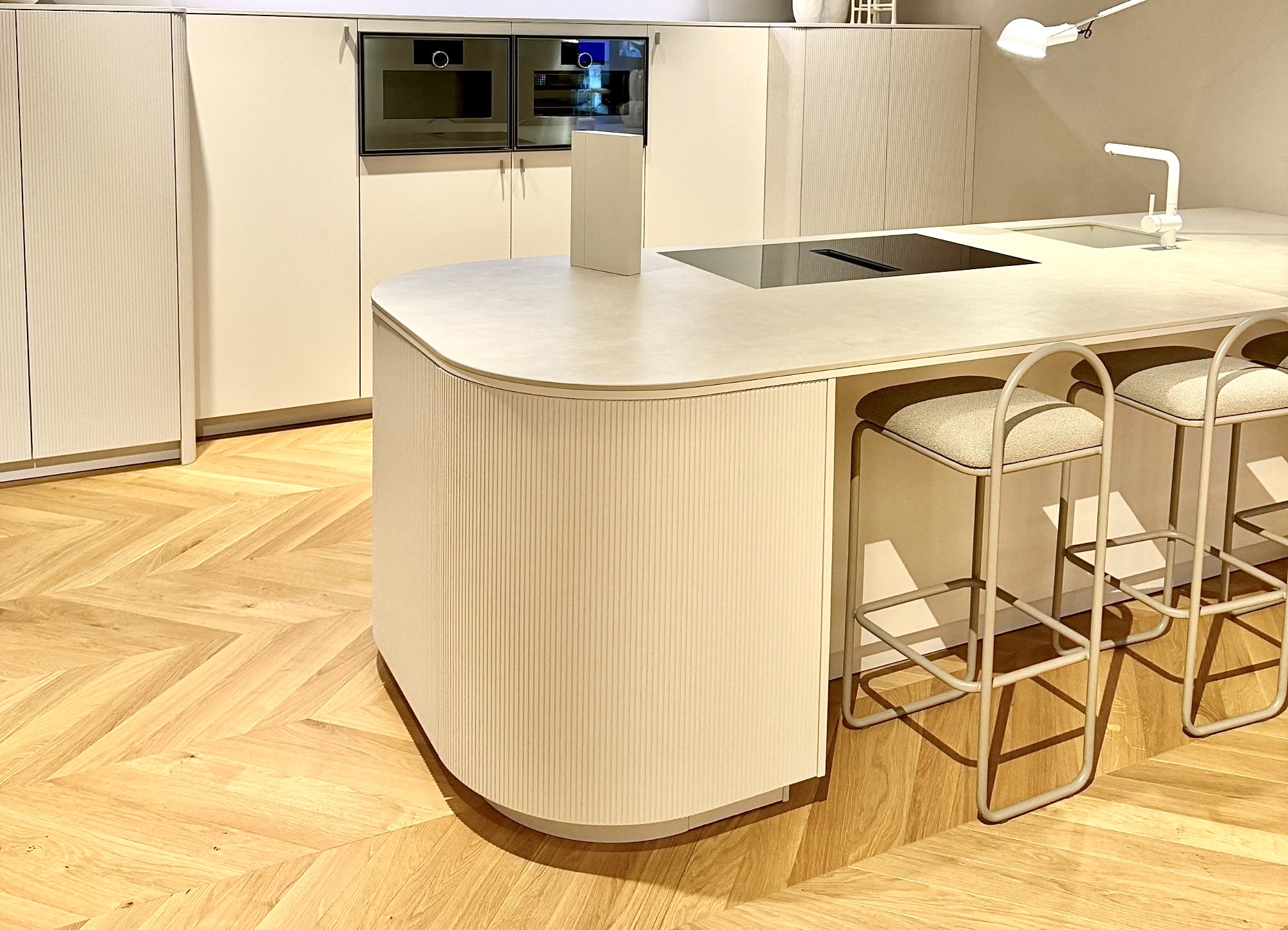 Helle Küche mit abgerundeter Kochinsel und Fronten in Rillenstruktur. Dezente Farben, grifflose Schränke und organische Formen setzen den Küchentrend 2026 für weiche, wohnliche Designs um.