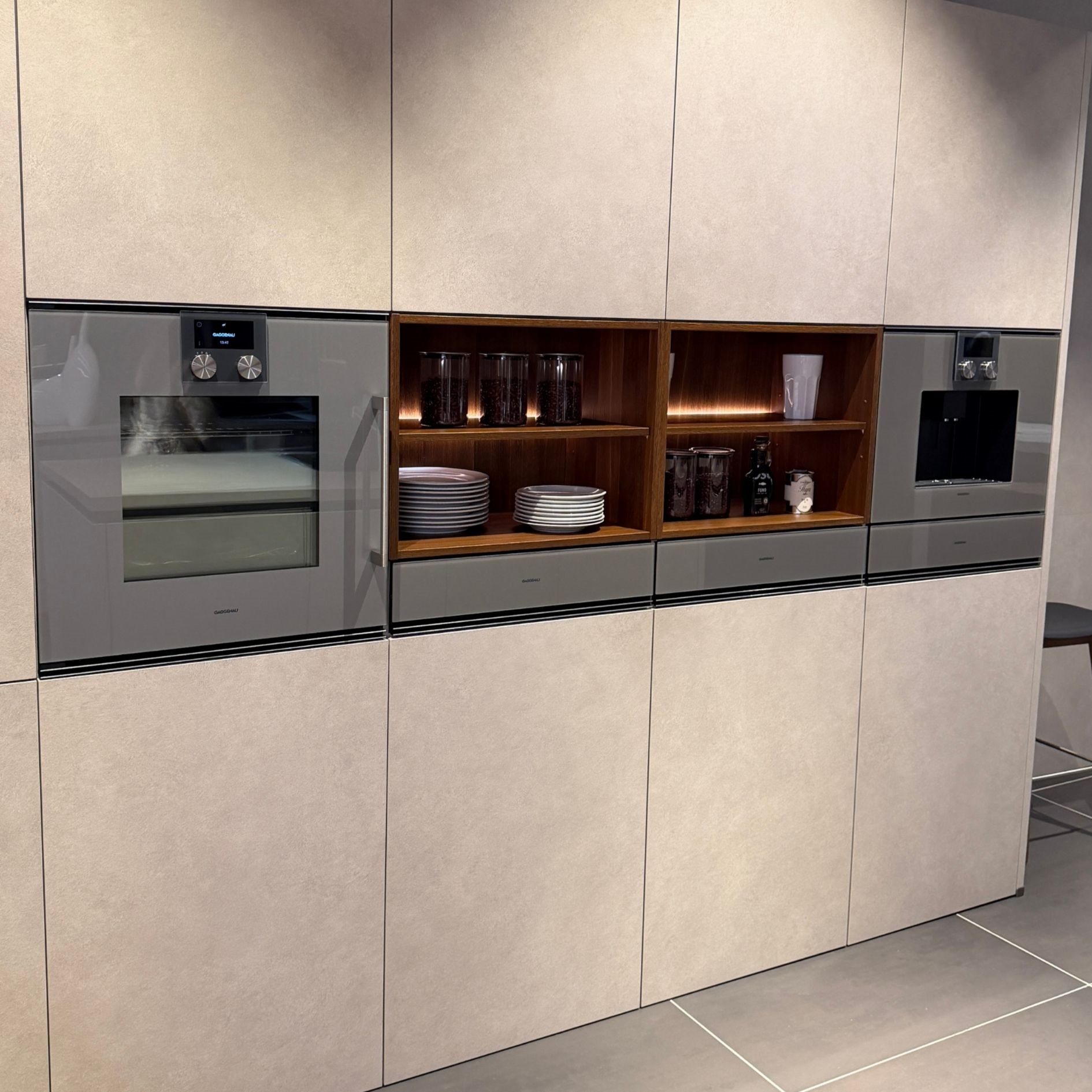 Eingebaute Küchengeräte in Grau-Matt mit offenen Holzregalen und Beleuchtung. Moderne Kombination aus Technik und warmen Materialien – ein Küchentrend 2026 für funktionales und stilvolles Design.