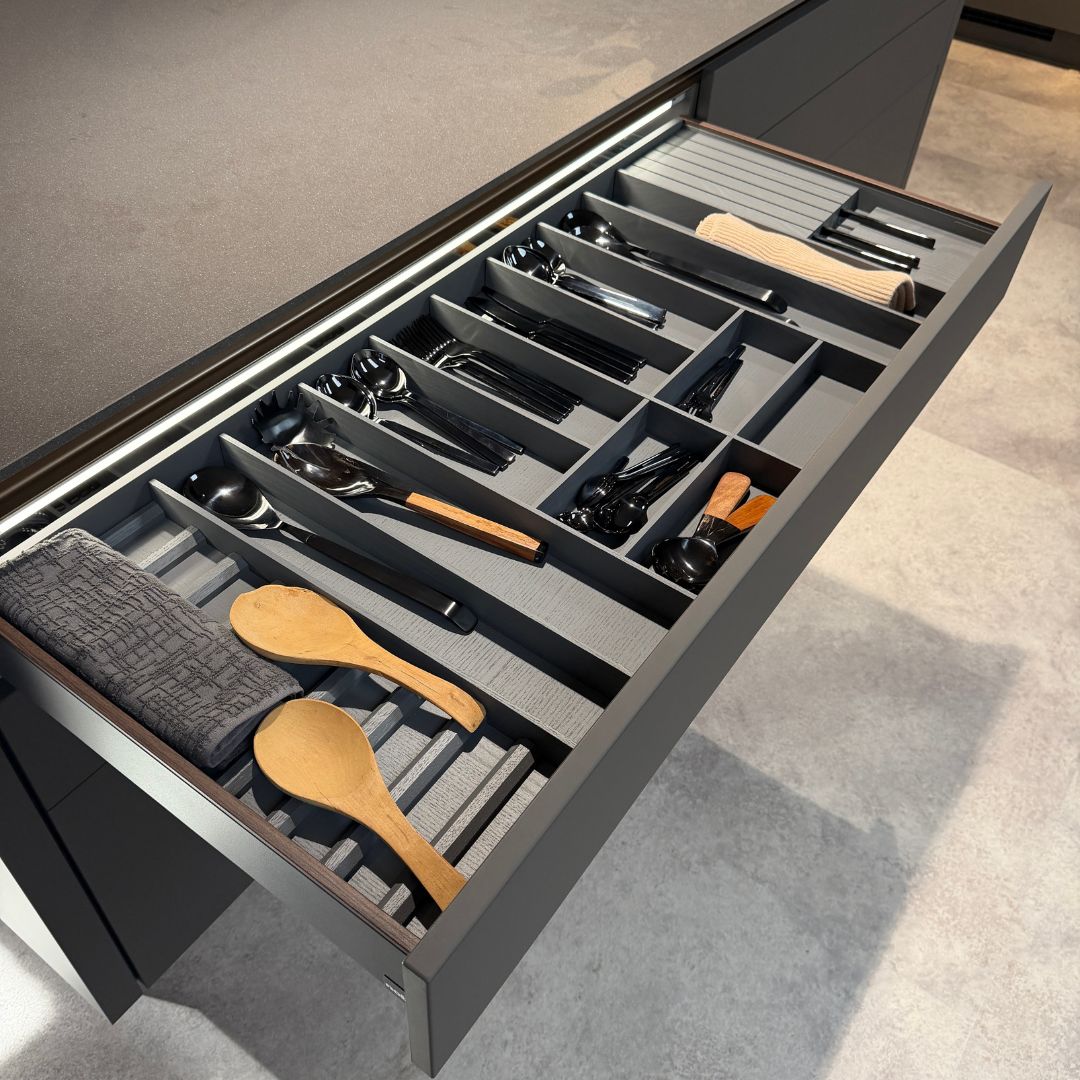 Offene Küchenschublade mit edlem Besteckeinsatz in Anthrazit und Holzoptik, organisiert mit schwarzem Besteck und Kochlöffeln, moderne Küche mit heller Arbeitsplatte und geradlinigem Design.