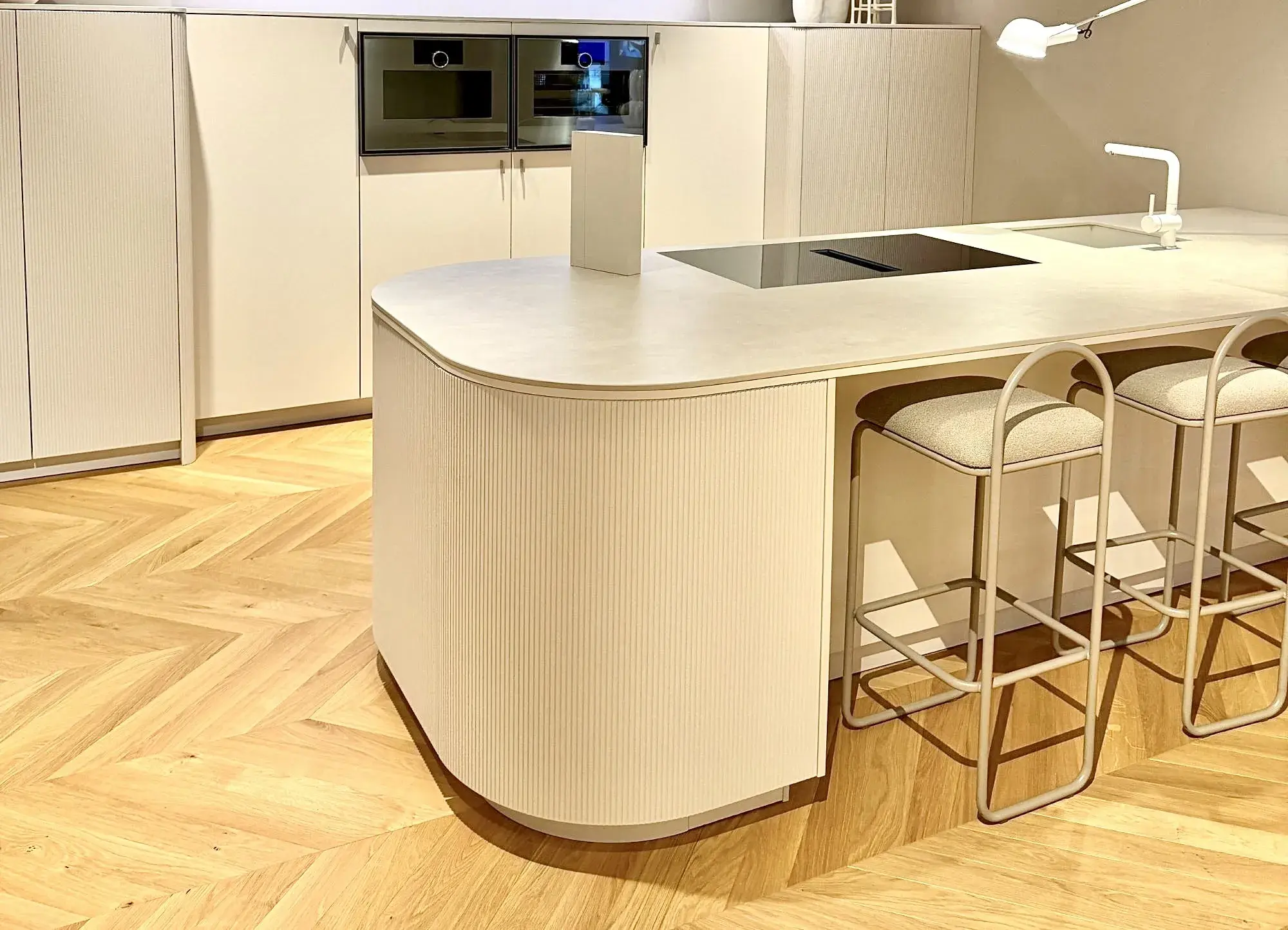 Helle Küche mit abgerundeter Kochinsel und Fronten in Rillenstruktur. Dezente Farben, grifflose Schränke und organische Formen setzen den Küchentrend 2026 für weiche, wohnliche Designs um.